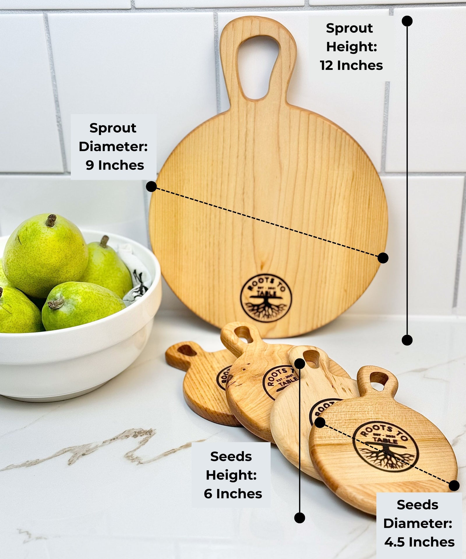 Round wooden cutting boards from Roots to Table, labeled with dimensions: Sprout board (12 inches tall, 9-inch diameter) and Seeds boards (6 inches tall, 4.5-inch diameter). Displayed on a white counter with pears in a bowl nearby.