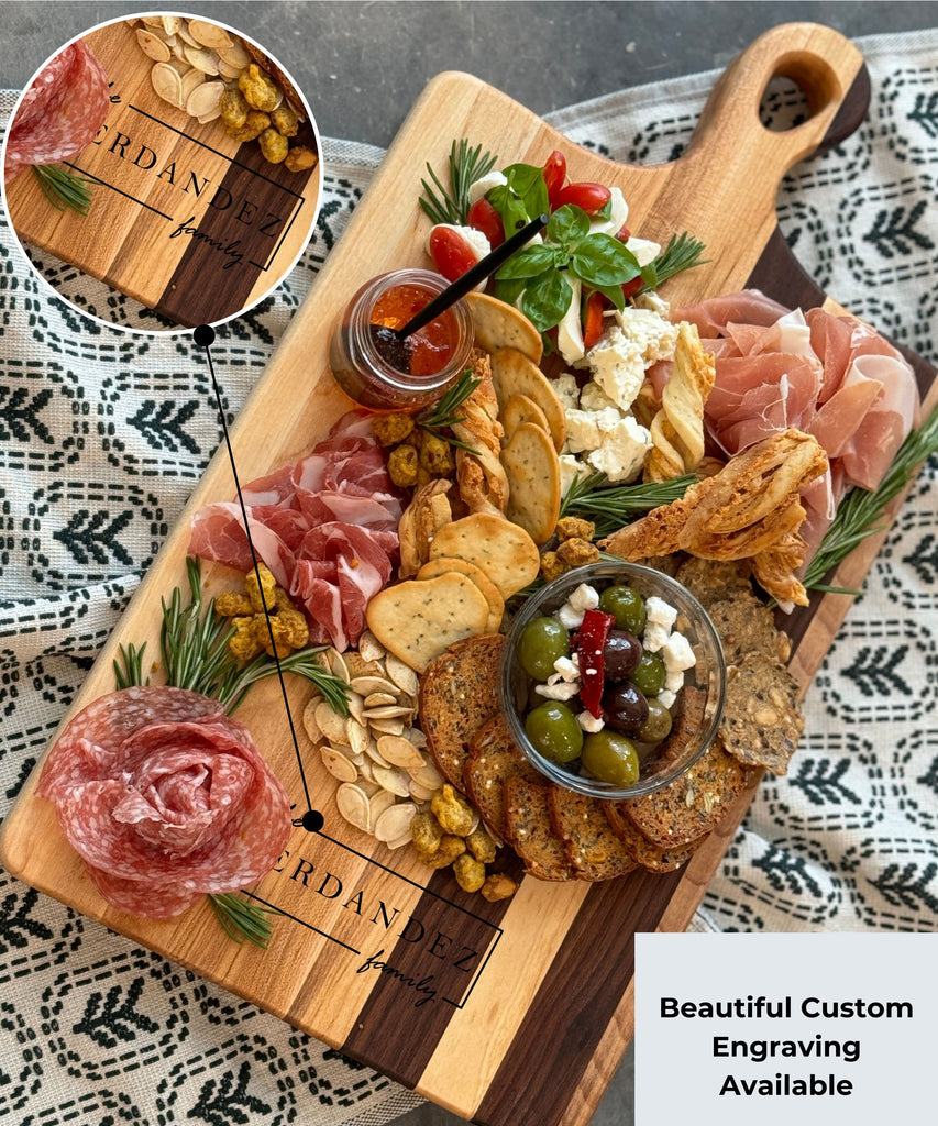 A personalized wooden charcuterie / cutting board featuring maple & walnut stripes, engraved with Fernandez Family. Topped fresh cherry tomatoes, olives with feta, herbs, & crispy crackers. Text overlay reads Beautiful Custom Engraving Available.