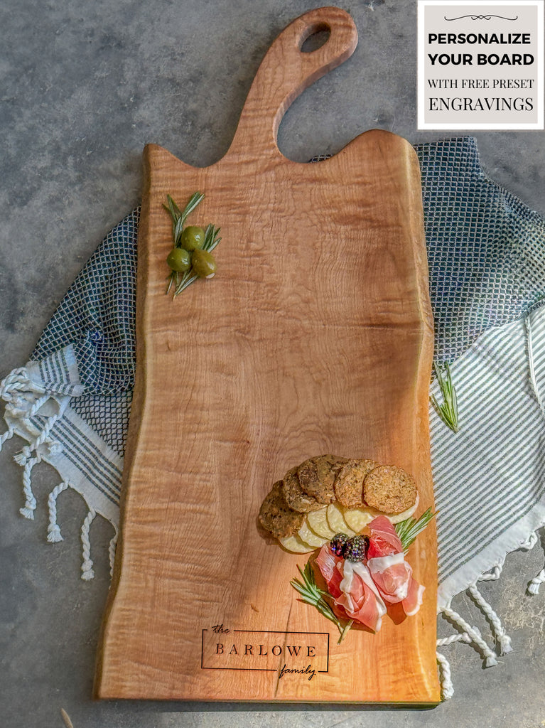 A personalized 24 inch maple live edge charcuterie board engraved with The Barlowe Family, featuring crackers, salami, cheese, and rosemary. The board rests on a striped, fringed towel against a concrete background.