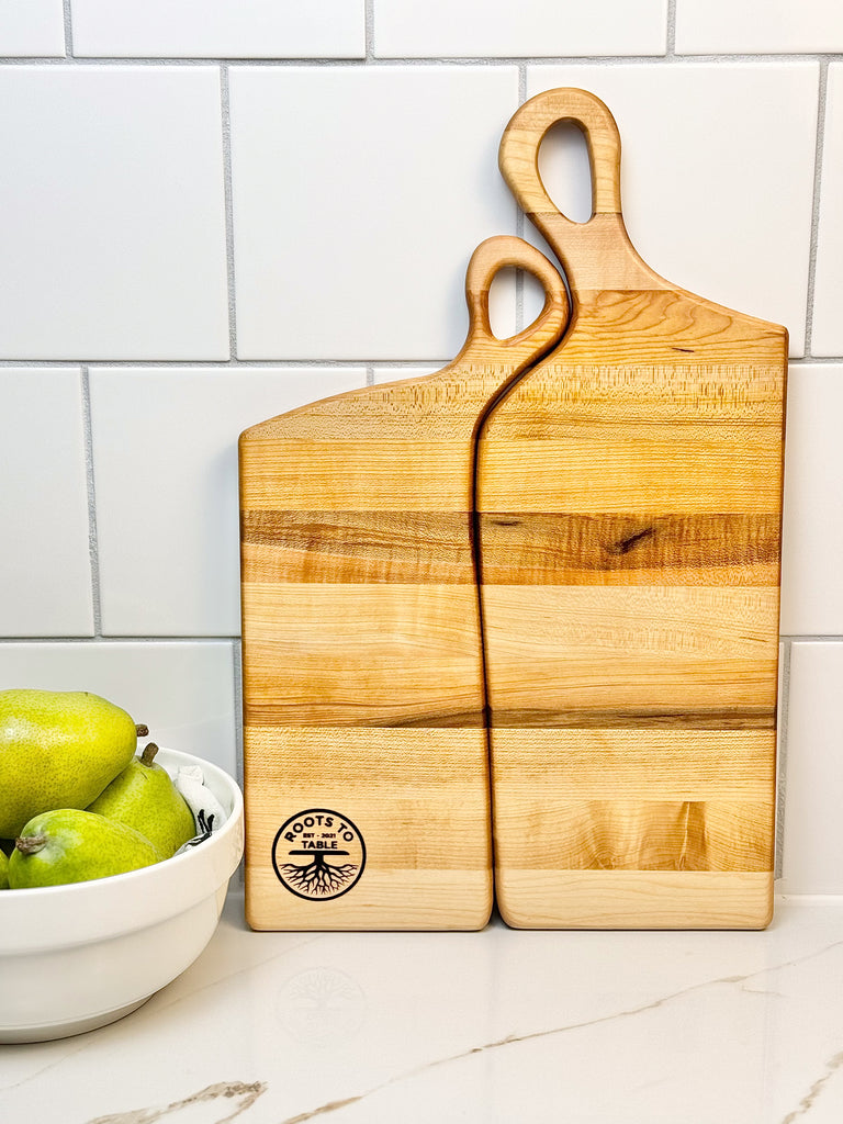 Two handcrafted wooden boards made from edge grain maple. Both feature ergonomic handles with a soft, rounded design, displayed on a textured fabric backdrop for a rustic aesthetic.