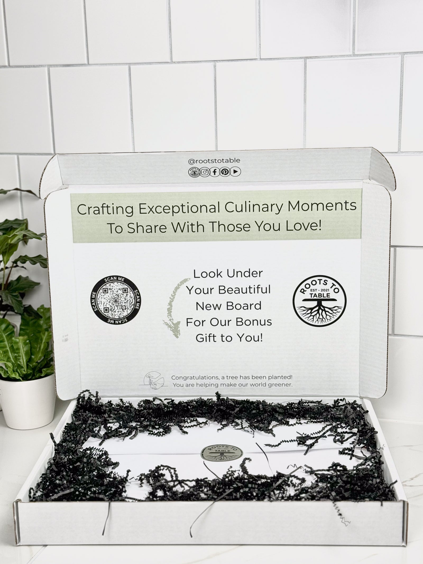 This image showcases how the charcuterie and cutting boards are packaged and presented as a gift It is wrapped in white paper held together with a logo sticker. Gaps are filled with black crinkle paper. Beautiful gift presentation