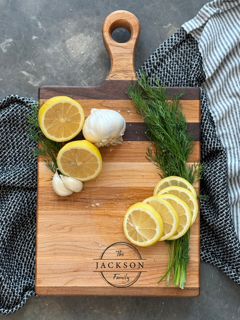 Small personalized wooden charcuterie / cutting board with The Jackson Family engraving. Featuring elegant walnut and maple stripes, garnished with Fresh dill, garlic cloves and sliced lemon. Perfect for hosting and gifting.