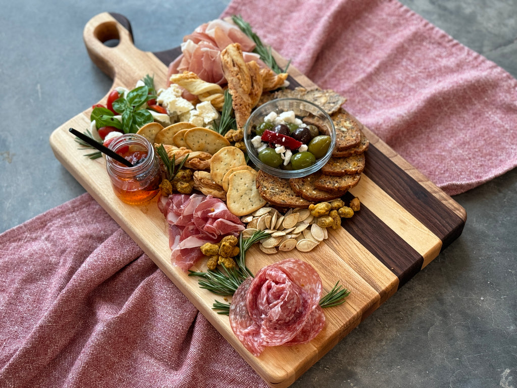 A beautifully arranged charcuterie board made of maple and walnut wood, featuring meats, cheeses, crackers, olives, and garnishes, set on a red fabric with a rustic gray background, showcasing the natural grain and craftsmanship of the board.