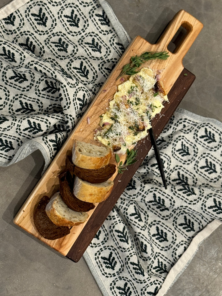 Using the backside of the the maple and walnut breadboard holding fresh bread slices and a creamy butter, garnished spread topped with herbs & parm cheese. Placed on a patterned cloth, creating a rustic yet elegant presentation.