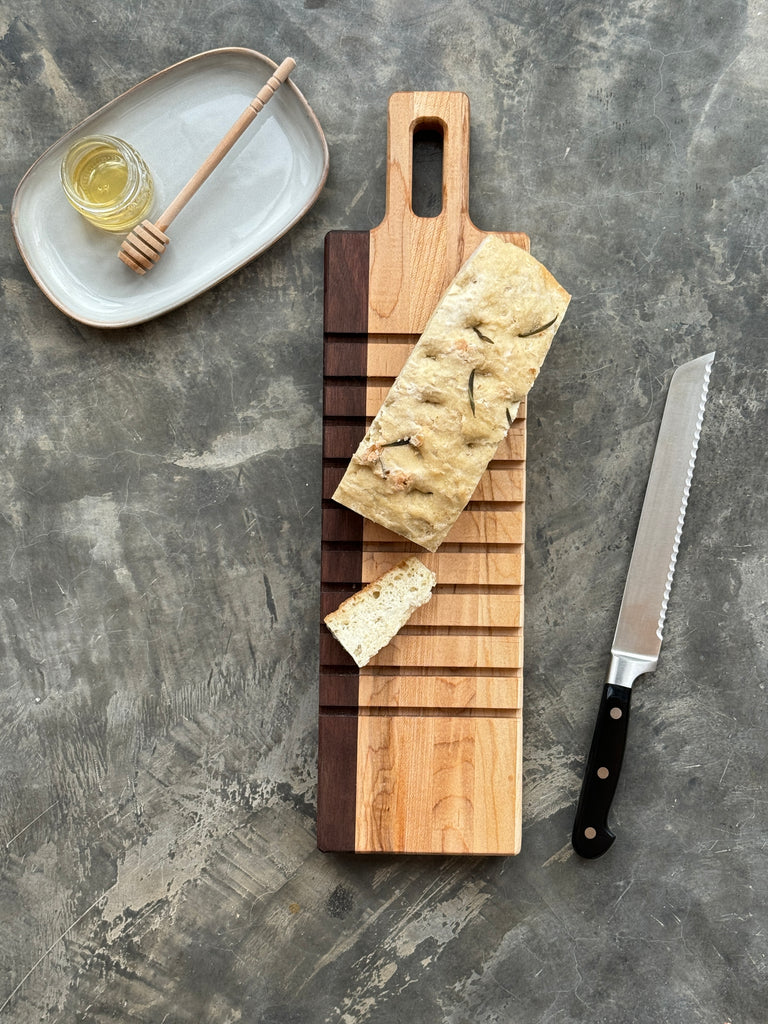 A wooden maple and walnut breadboard for slicing bread is featured showing a loaf of bread, a bread knife and honey and dipper.