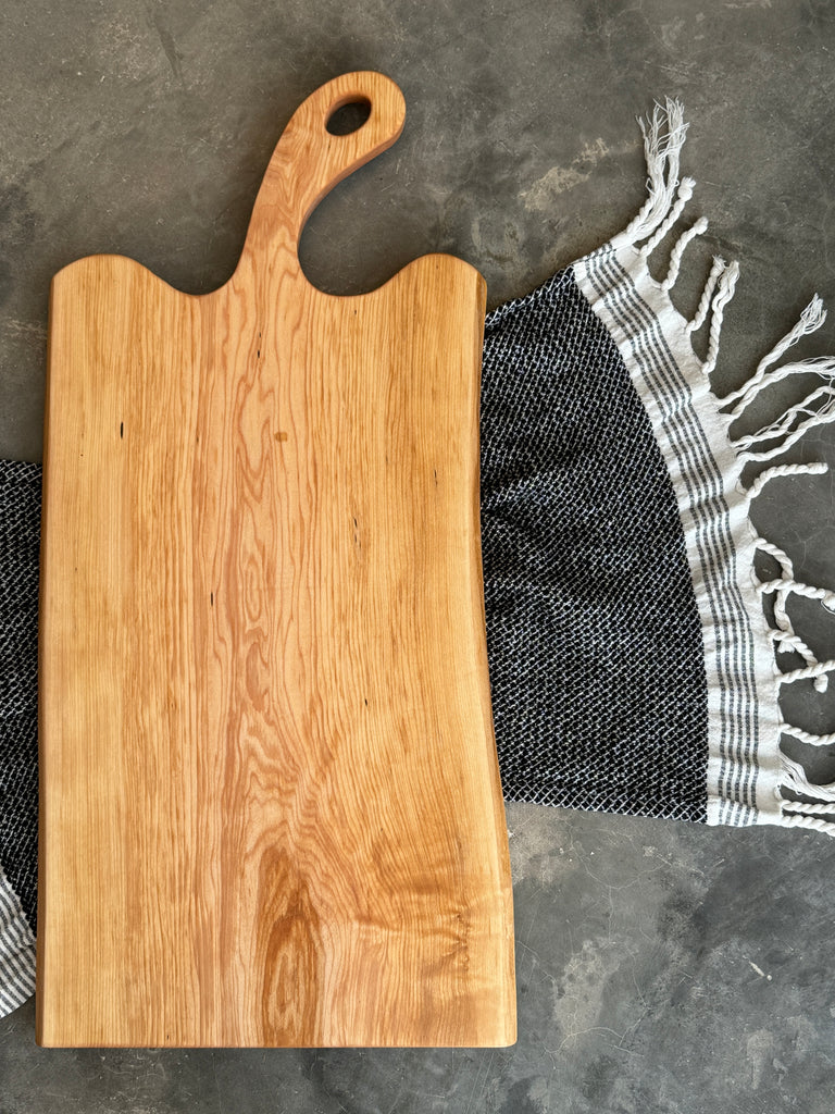 A handcrafted face grain maple wooden charcuterie board with a curved handle, made from beautifully grained light wood. The board has a natural, organic shape and is placed on a textured surface with a black and white striped fringed cloth partially underneath.