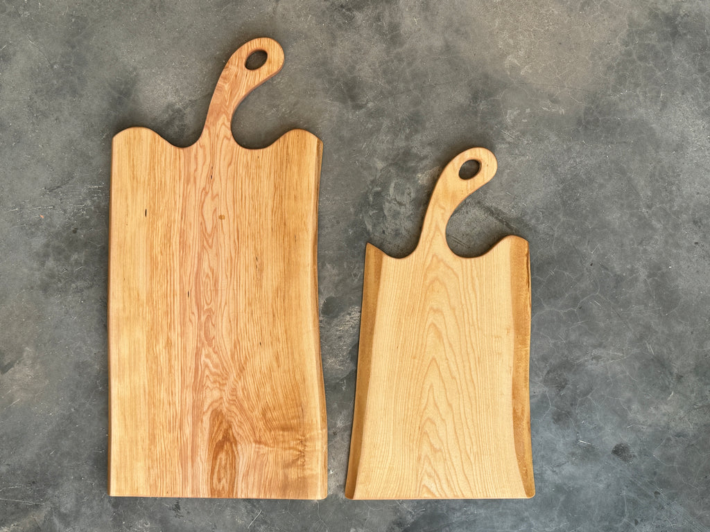Two handcrafted face grain maple wooden charcuterie boards with a curved handles, one is 18inch long, one is 24inch. The board has a natural, organic shape and is placed on a textured surface with a black and white striped fringed cloth partially underneath.