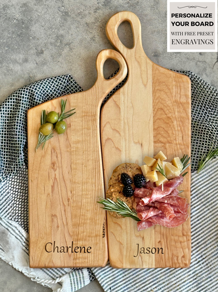 Two handcrafted wooden boards made from edge grain maple. Both feature ergonomic handles with a soft, rounded design, displayed on a textured fabric backdrop for a rustic aesthetic.