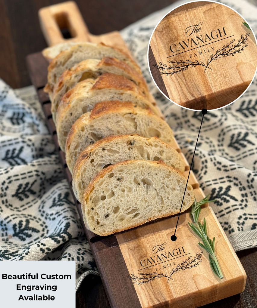 A beautifully crafted breadboard featuring The Cavanagh Family engraving with a delicate laurel design. Slices of fresh bread rest on the board, paired with sprigs of rosemary, set against a patterned cloth backdrop. Perfect for elegant serving.