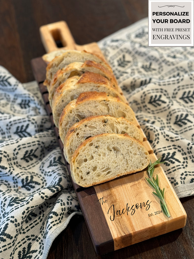 A beautifully crafted walnut breadboard featuring The Jacksons engraving with established date. Slices of fresh bread rest on the board, paired with sprigs of rosemary, set against a patterned cloth backdrop. Perfect for elegant serving.