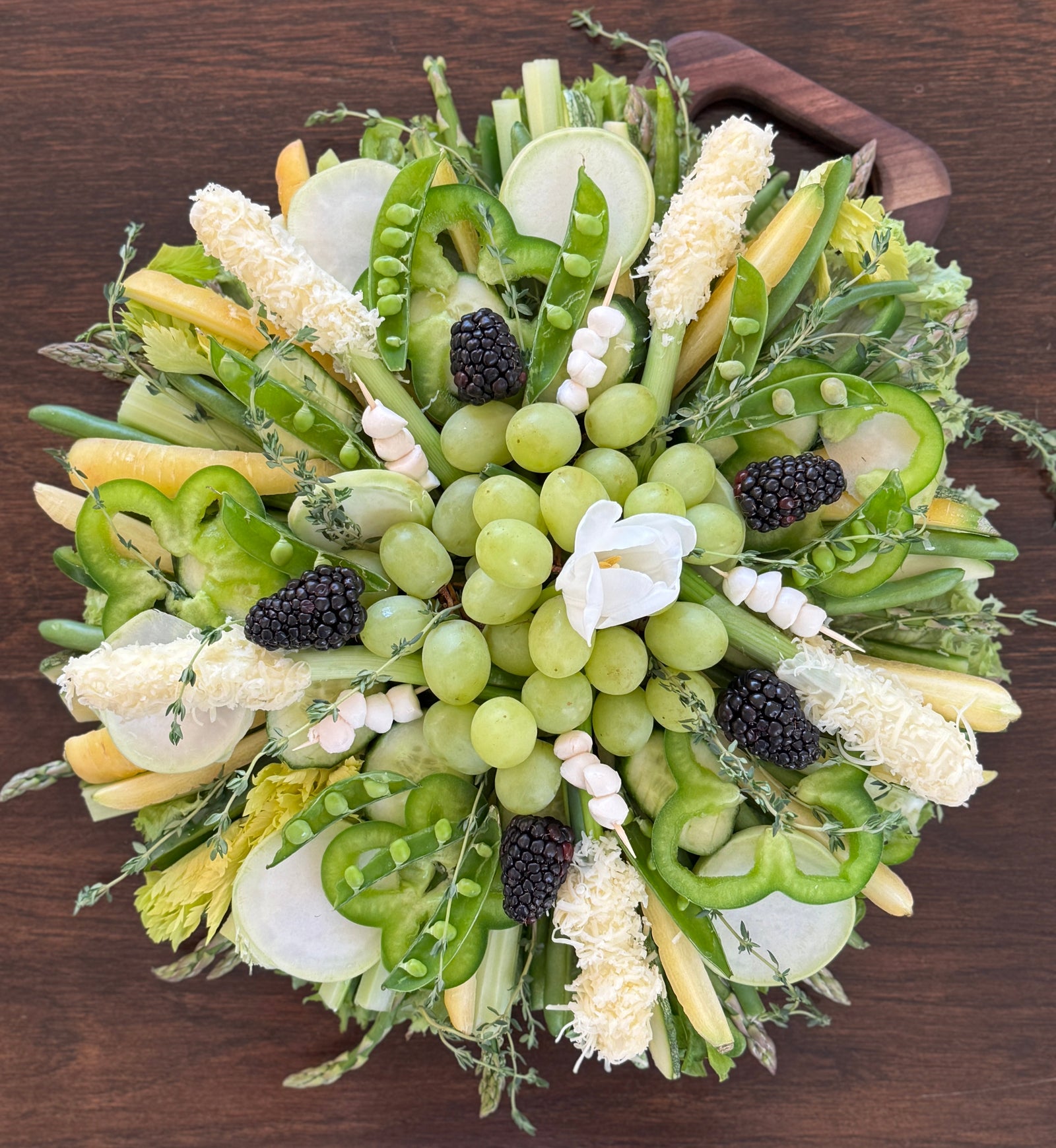 a stunning circular shaped charcuterie board displaying carrots, celery, green pepper, snap peas, asparagus, zucchini, grapes, guava and kohlrabi.