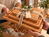 A set of three stacked solid  quality maple wooden cutting boards with a burlap ribbon and a paper tag reading 'The Bakers', being held by people in a warm, rustic kitchen setting.