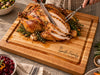 A roasted turkey on a large personalized wooden cutting board, with a knife slicing the breast and fork holding a cut piece. The board has the text The Smith Family Est 2026 engraved on the front, and holiday dishes are arranged around it on a stone surface.