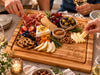 A wooden charcuterie board engraved with 'Meat you at The Millers' is styled on a dining table with hands reaching in to grab food, including sliced cheese, crackers, fruit, nuts, and cured meats. Wine glasses and candles are visible around the table.