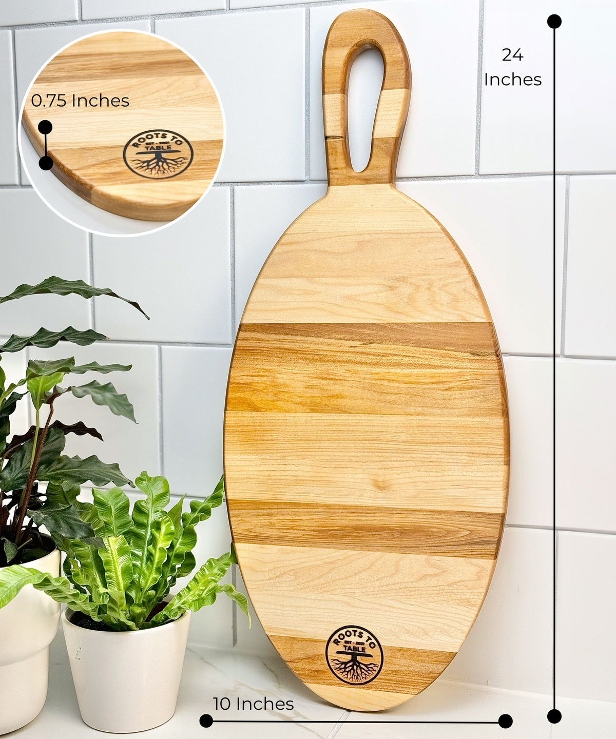  An oval-shaped 24-inch Canadian maple charcuterie board with a handle, featuring a Roots to Table logo. Highlighted features include beautiful craftsmanship, mineral oil treatment for a glossy finish, and ample space for entertaining. Displayed against a tiled wall with a green plant nearby.