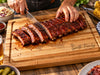 A close-up of a person slicing a cooked rack of ribs on a personalized wooden chopping board with engraved script reading 'The Smith Family Est. 2024.