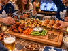 A large wooden serving board at a social gathering, loaded with buffalo wings, nachos, celery, carrots, dip, and sliders. Several people are visible around the table, wearing Toronto Maple Leafs sweaters and holding beer bottles and glasses, with a TV in the background.