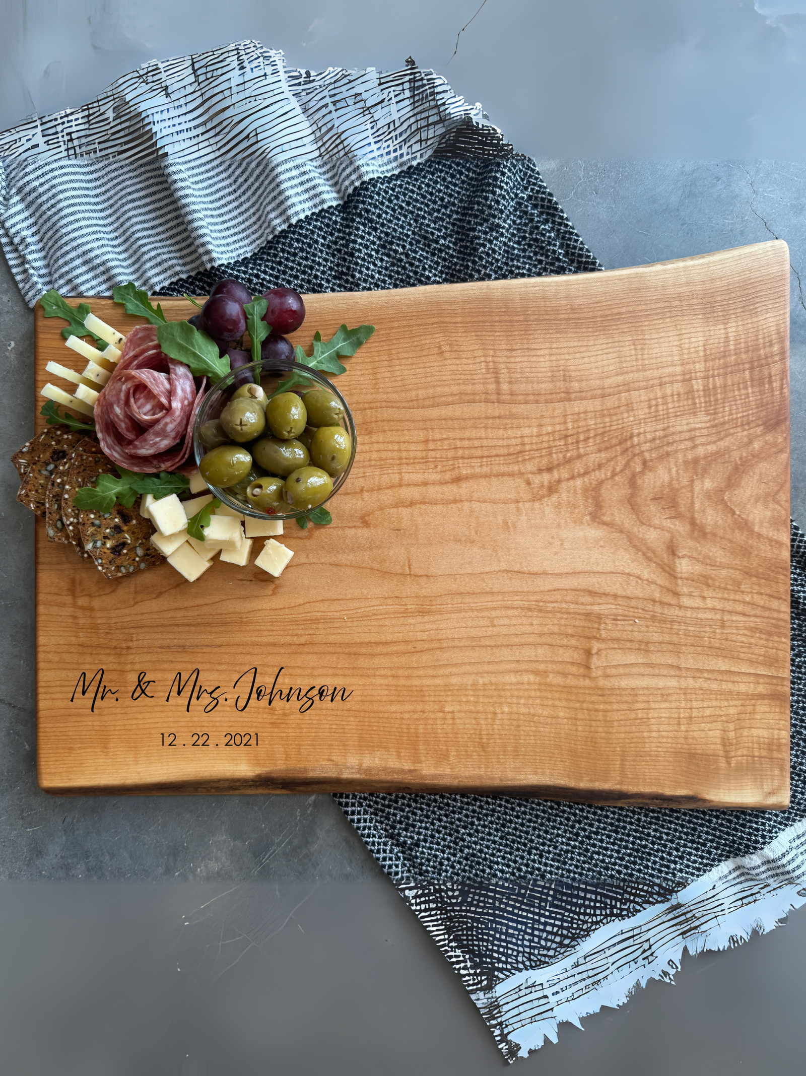 A maple charcuterie board with a natural live edge. The board is styled with a small arrangement of olives, crackers, cheese cubes, grapes, and salami at the top. A black and white striped cloth is partially visible underneath, with a 'Personalize Your Board' engraving promotion displayed in the corner."
