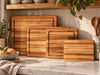 Four maple wooden cutting boards of different sizes arranged on a kitchen counter, with a marble surface, tiled backsplash, wooden shelf, and kitchen props including lemons, a bottle, and utensils. The smallest board is engraved with the brand name Holly's Kitchen.