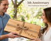 A man and a woman outdoors holding a personalized wooden gift box tied with twine. The box has a live edge, natural wood grain, and is engraved with 'The Browns EST. Wood is the traditional 5th wedding anniversary gift.