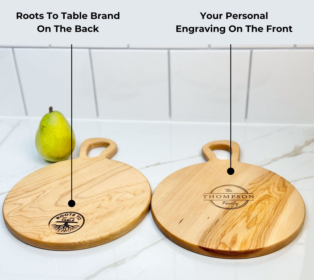 Two round wooden cutting boards from Roots to Table: one displays the brand logo on the back, while the other showcases a custom engraving reading The Thompson Family on the front. A green pear is placed nearby on a white countertop.