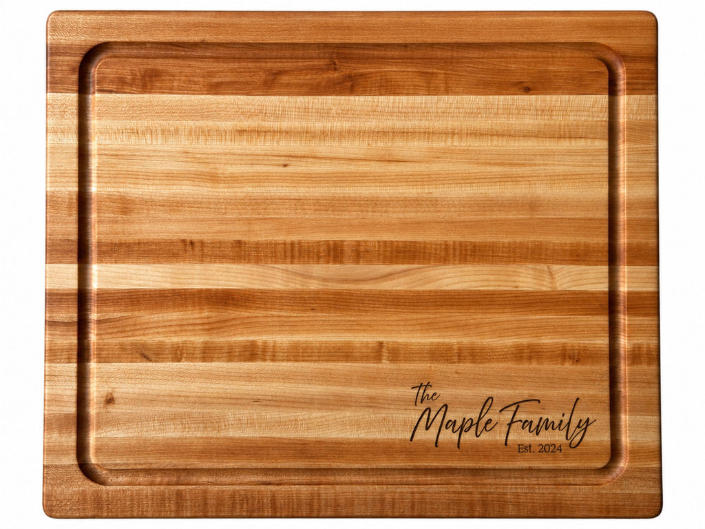 An extra large rectangular wooden cutting board with a raised rim and channeled edges for a juice groove, featuring a natural maple honey-toned wood grain and an engraved script logo reading The Maple Family Est. 2024 in the bottom right corner.