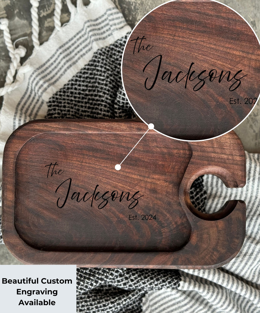 A Dark walnut wood wine appetizer tray with a custom engraving that reads The Jacksons, Est. 2024, placed on a black and white patterned cloth. The tray features a circular cutout for holding a wine glass, showcasing elegant personalized design options.