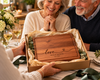 Smiling older couple receiving a handcrafted personalized wooden cutting board presented in a gift box with natural tissue paper. The board features a custom engraved message and warm cherry wood grain. A thoughtful gift for parents, grandparents, wedding anniversaries, Christmas or special family celebrations
