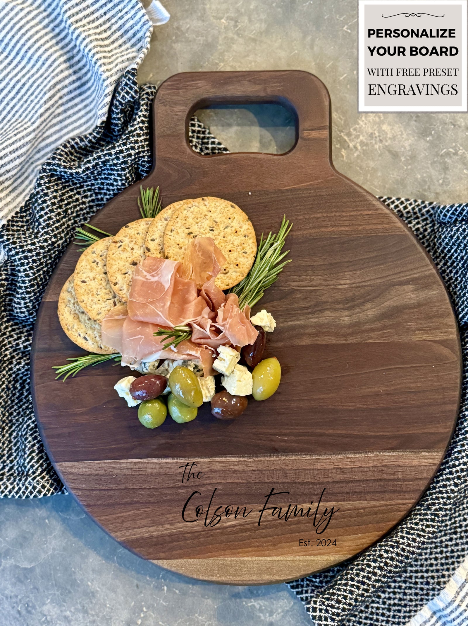 Round wooden charcuterie board with a smooth walnut finish featuring customizable engraving for Olivia & Alex, 12.22.2021. Decorated with neatly arranged salami, cheese, and herbs, perfect for gifting or entertaining. Background includes greenery and textiles.