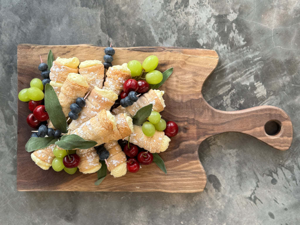 A handcrafted, solid walnut live edge charcuterie board featuring rich natural wood grain, a curved handle with a rounded cutout. Cannoli filled with custard, cherries, grapes and blueberries are displayed atop the board.