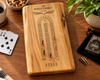 A personalized maple wooden cribbage board engraved with 'The White Family Est. 2021' and a cribbage track, styled on a wooden table with playing cards, a coffee cup, nuts, a plant, and a black Bicycle Spades card deck.
