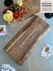 A live edge walnut solid wood cribbage board engraved with The Smith Family, Est. 1980, displayed alongside a live edge walnut charcuterie spread with cheese, meats, and olives. Cards and a wine glass complete the cozy, personalized game night setup