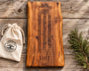 A live-edge wooden crib board game set on a rustic wood plank background, featuring a dark brown engraved board with a curved track and a small cream drawstring pouch printed with 'ROOTS TO TABLES' on the left, and a green pine branch on the right.
