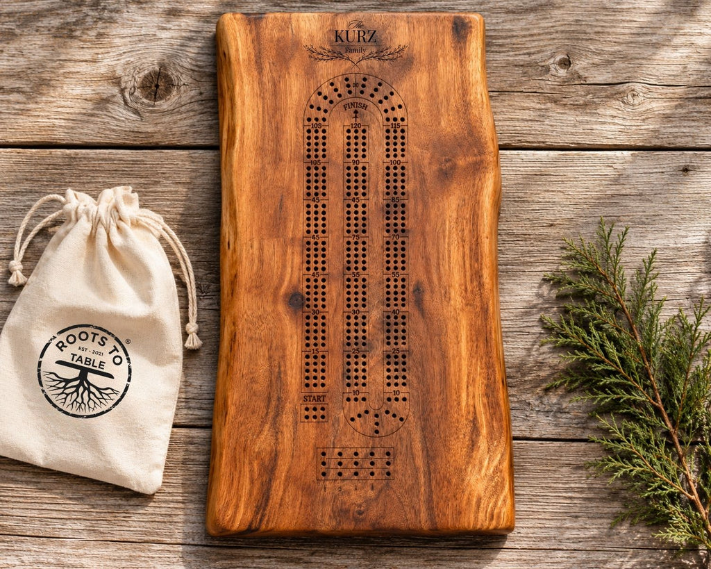 A live-edge wooden crib board game set on a rustic wood plank background, featuring a dark brown engraved board with a curved track and a small cream drawstring pouch printed with 'ROOTS TO TABLES' on the left, and a green pine branch on the right.