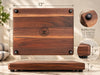 Personalized hardwood cutting board measuring 12 inches by 9 inches, featuring four black rubber feet underneath for added stability and countertop clearance. Detailed product image highlighting the board's thickness, natural wood construction, routed juice groove and non-slip feet. A compact handcrafted cutting board for everyday kitchen use.