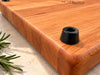 Close-up of a handcrafted wooden cutting board showing its smooth rounded edges, natural wood grain and small black rubber foot. The non-slip rubber feet are attached underneath the board to provide stability and elevate it slightly above the countertop. A practical detail of this premium personalized hardwood cutting board.