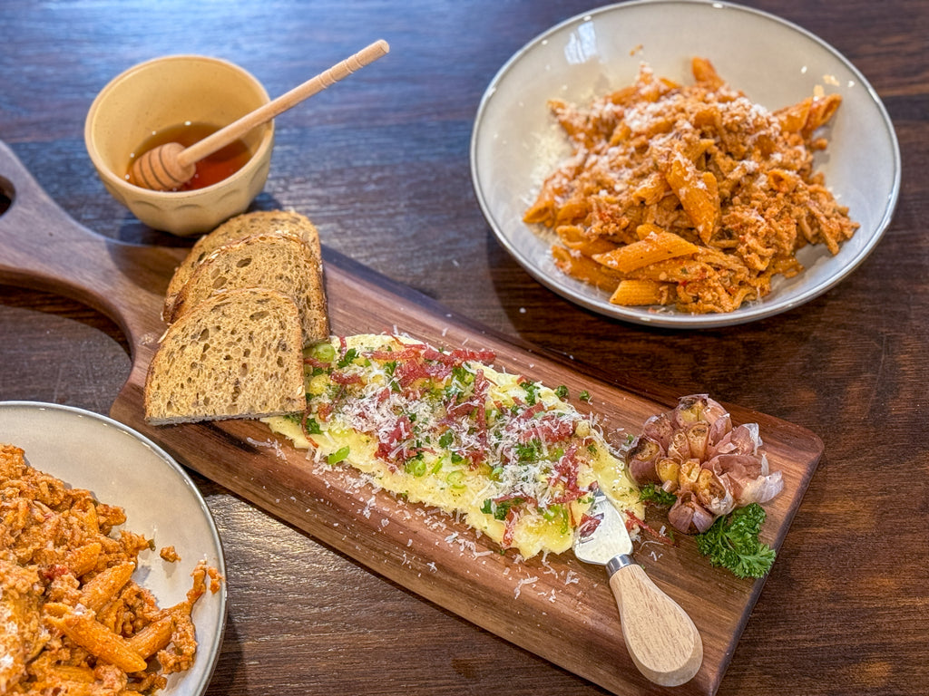 Two bowls of pasta atop a table with a small black walnut live edge charcuterie sits in the centre. The board as a butter spread and fresh cut bread with a large bulb of roasted garlic.