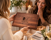 Woman surprising her friend with a handcrafted personalized walnut cutting board featuring an engraved illustration of two best friends and the message We're more than friends, we're like a really small gang. A thoughtful friendship gift presented in a cozy home setting, perfect for birthdays, Christmas or celebrating a special friendship.