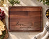 Overhead view of a handcrafted walnut wood cutting board featuring a custom engraved love design, Jenna and Mick, Est. 2026. Rich natural walnut grain, rounded corners and a precision-routed juice groove are showcased against a warm neutral countertop. A personalized wooden cutting board for wedding, anniversary and housewarming gifts.