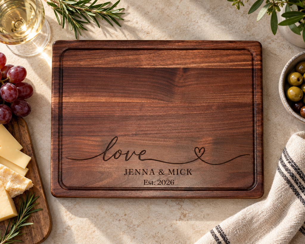 Overhead view of a handcrafted walnut wood cutting board featuring a custom engraved love design, Jenna and Mick, Est. 2026. Rich natural walnut grain, rounded corners and a precision-routed juice groove are showcased against a warm neutral countertop. A personalized wooden cutting board for wedding, anniversary and housewarming gifts.