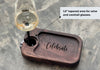 Dark walnut wine and appetizer tray featuring an engraved, Celebrate, text, with a tapered stemmed glass holder designed for wine or cocktail glasses, placed on a rustic concrete surface