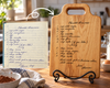 A wooden cutting board with a handle stands on a black metal easel in a kitchen setting, displaying a custom chocolate brownie recipe engraved on it. An inset shows a handwritten recipe note labeled 'Chocolate Brownie' with a white arrow pointing to the board demonstrating how the handwritten recipe is engraved exactly as is onto the board.