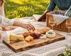 A wooden charcuterie board engraved with 'Home Sweet Home' is set on a white crochet picnic blanket outdoors. The board is styled with cheese wedges, brie, a baguette, red grapes, and rosemary, with wine glasses, a basket, and people's hands and arms visible in a sunny grassy park setting.