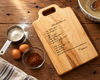 A handwritten recipe memorialized on a maple wooden cutting board with a handle, engraved with a recipe titled Chocolate Brownies, styled on a rustic wooden table with baking ingredients including flour, eggs, cocoa powder, and a bowl of batter.