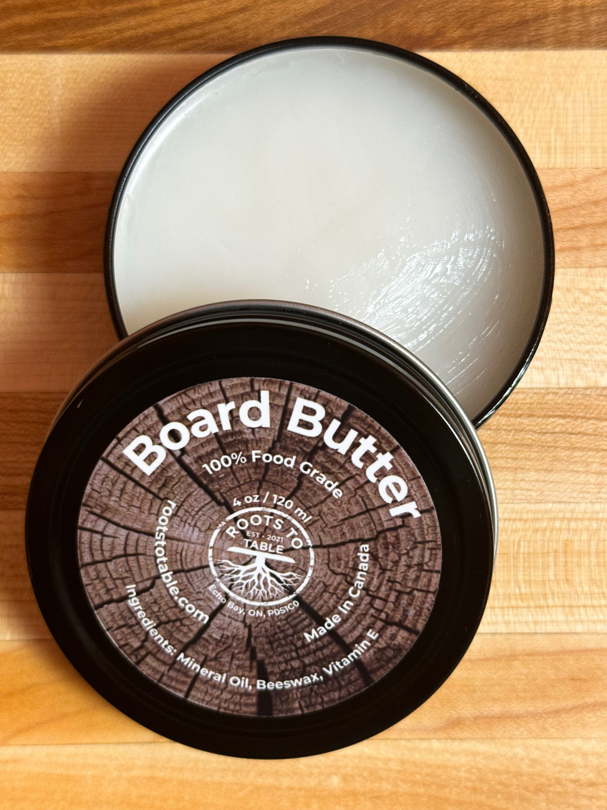 Tin of Roots to Table Board Butter, labeled as 100% food grade with ingredients including mineral oil, beeswax, and vitamin E. The product is displayed on a wooden surface with a "Pack of 4" label highlighted. Made in Canada, 4 oz/120 ml size.