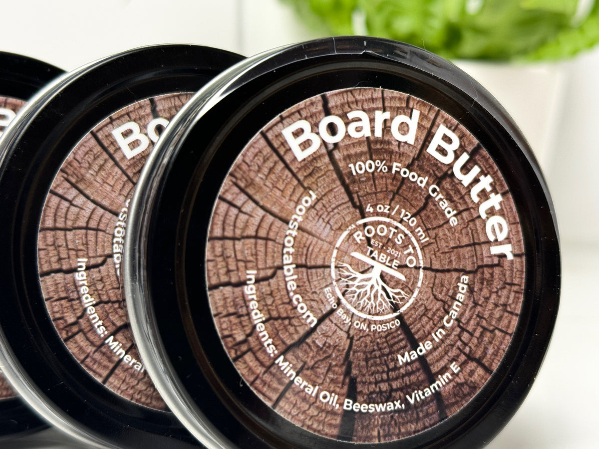 Tin of Roots to Table Board Butter, labeled as 100% food grade with ingredients including mineral oil, beeswax, and vitamin E. The product is displayed on a wooden surface with a "Pack of 4" label highlighted. Made in Canada, 4 oz/120 ml size.