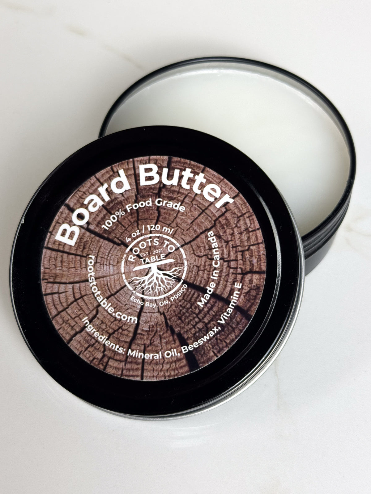 An open Tin of Roots to Table Board Butter, labeled as 100% food grade with ingredients including mineral oil, beeswax, and vitamin E. 