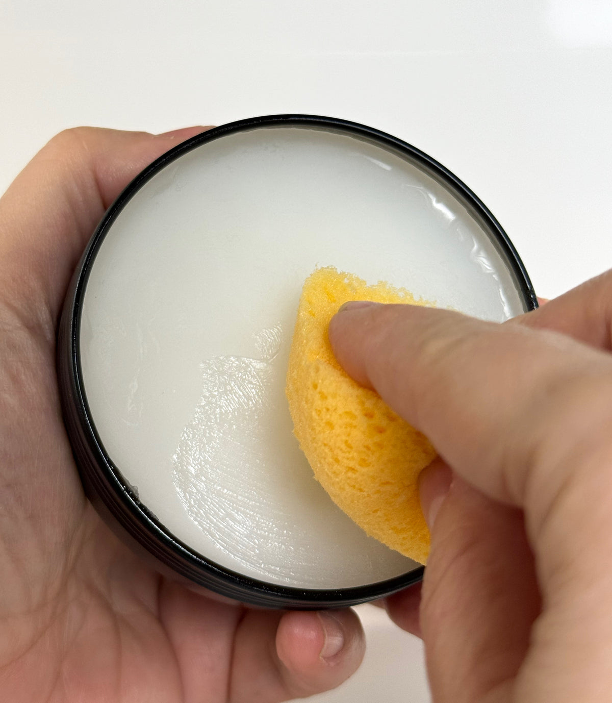 A person using a yellow sponge to scoop Roots to Table Board Butter from a tin, showcasing the creamy consistency of the food-safe finish. The background is clean and white, emphasizing the product application.