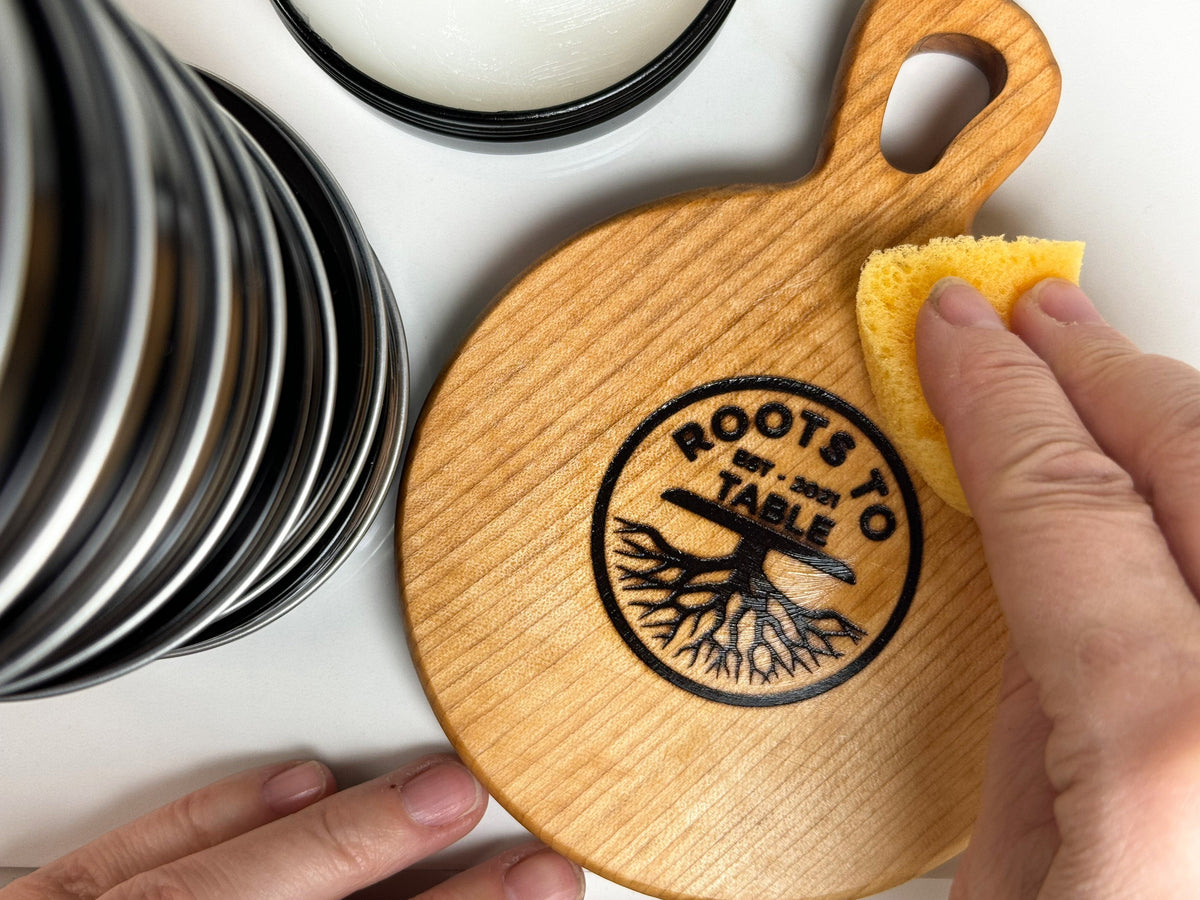 A person using a yellow sponge to apply Roots to Table Board Butter from a tin, to a maple board. 