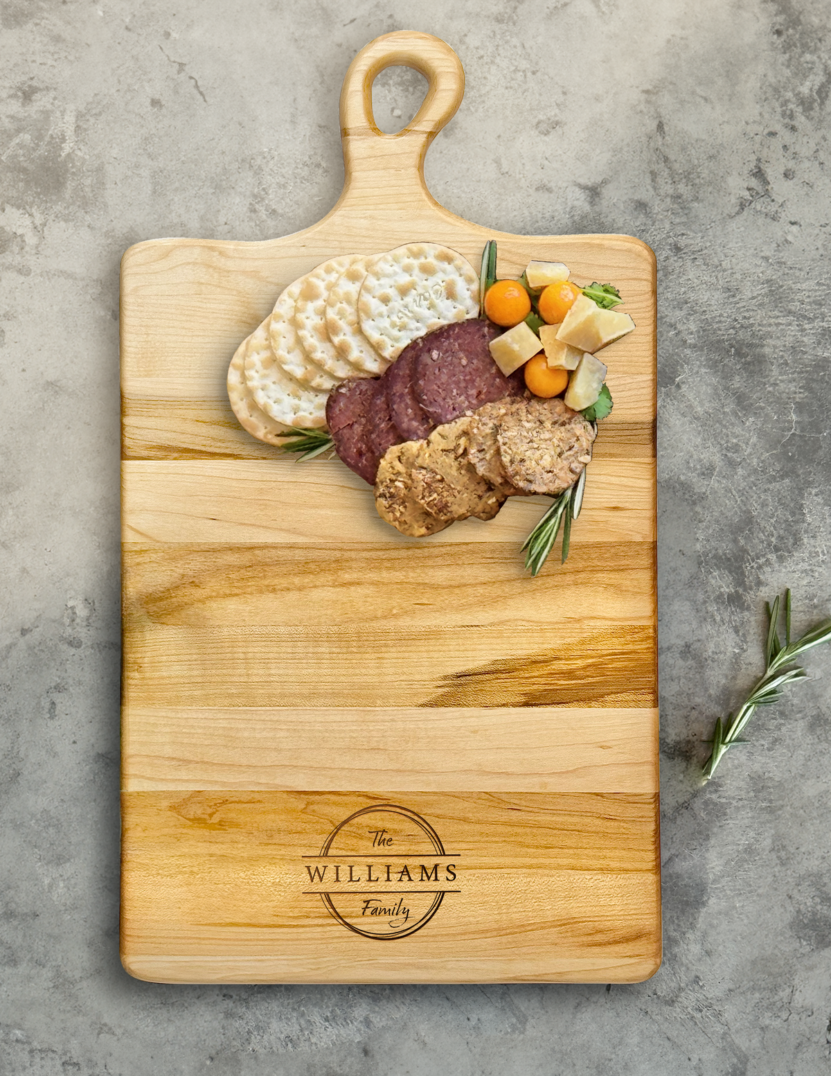 A personalized maple wood charcuterie board with The Williams Family engraving, beautifully arranged with cheese set on a concrete countertop.
